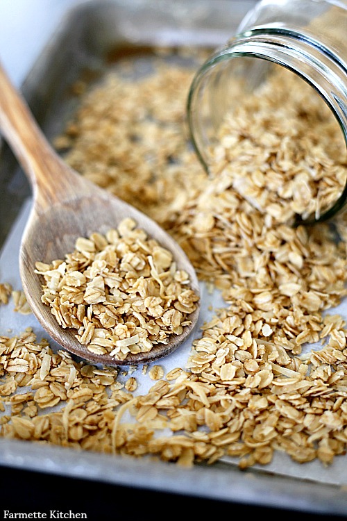 granola spilling out of a jar onto a wooden spoon and baking sheet