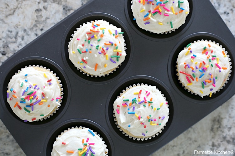 six cupcakes in a muffin tin with vanilla buttercream frosting and sprinkles